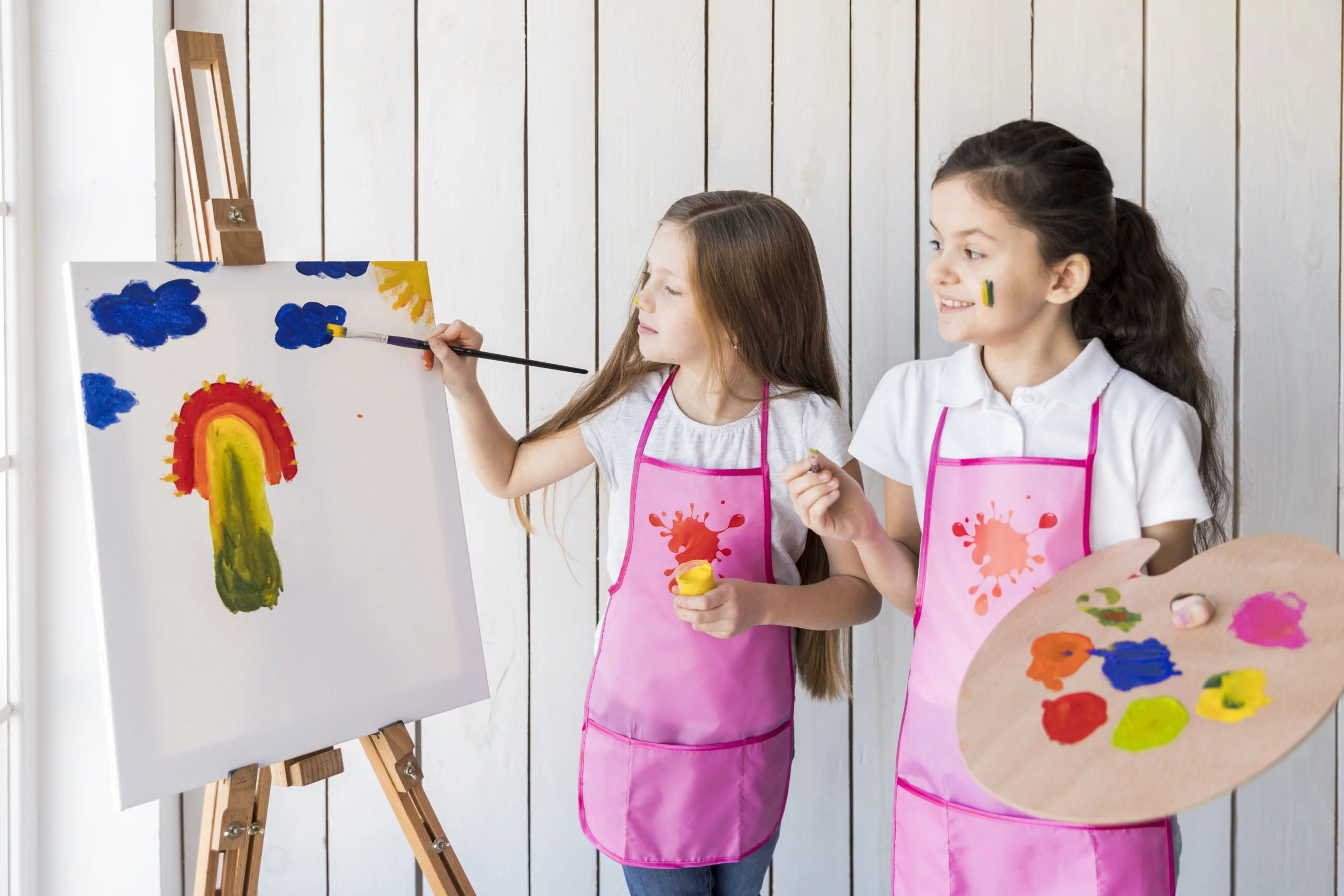 happy-girl-holding-palette-hand-looking-her-friend-painting-easel-with-paint-brush