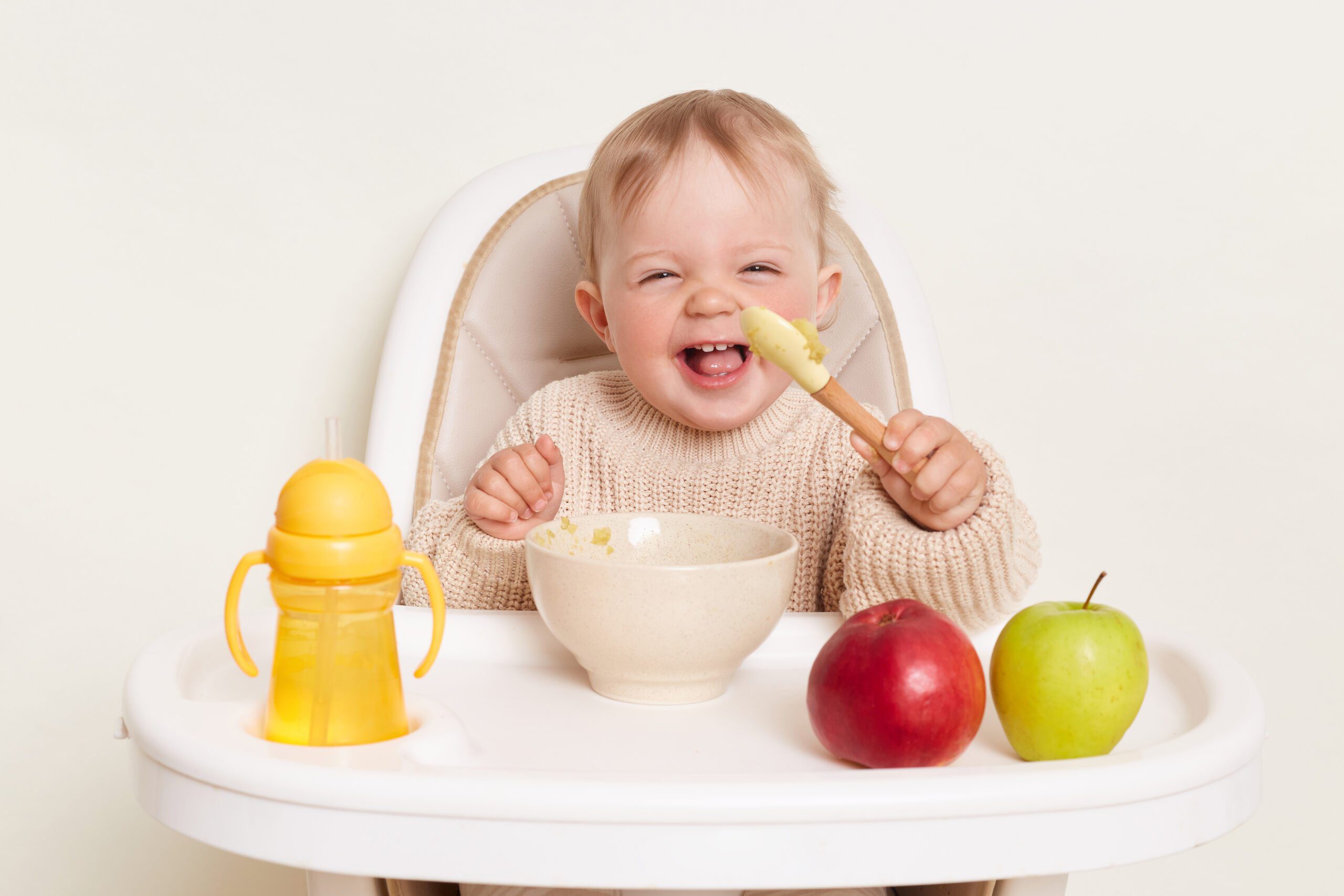 image-happy-positive-baby-wearing-beige-sweater-sitting-child-s-chair-eating-puree-laughing-holding-spoon-posing-alone-isolated-white-background